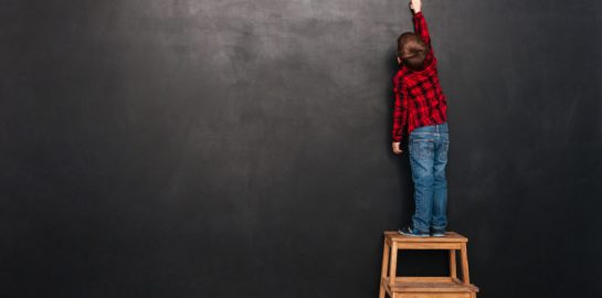 Picture of a little child standing on stool near blackboard and drawing on it.